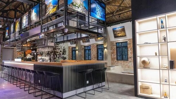 Un área de bar moderna con una barra larga y varios taburetes, una pared de ladrillo con un mural, y una exhibición de diversos objetos en estanterías.