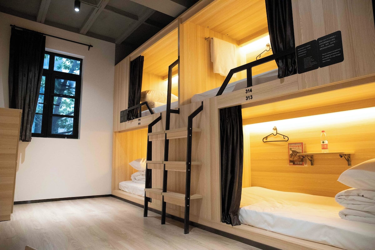 Habitación con literas de madera y ventana. Una escalera conecta las camas, y hay un estante con una botella y un libro.
