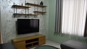 A living room with a flat-screen TV, wooden shelves, a sofa, a coffee table, and a window with curtains. - Modern Apartment In City Center (Tiranë)