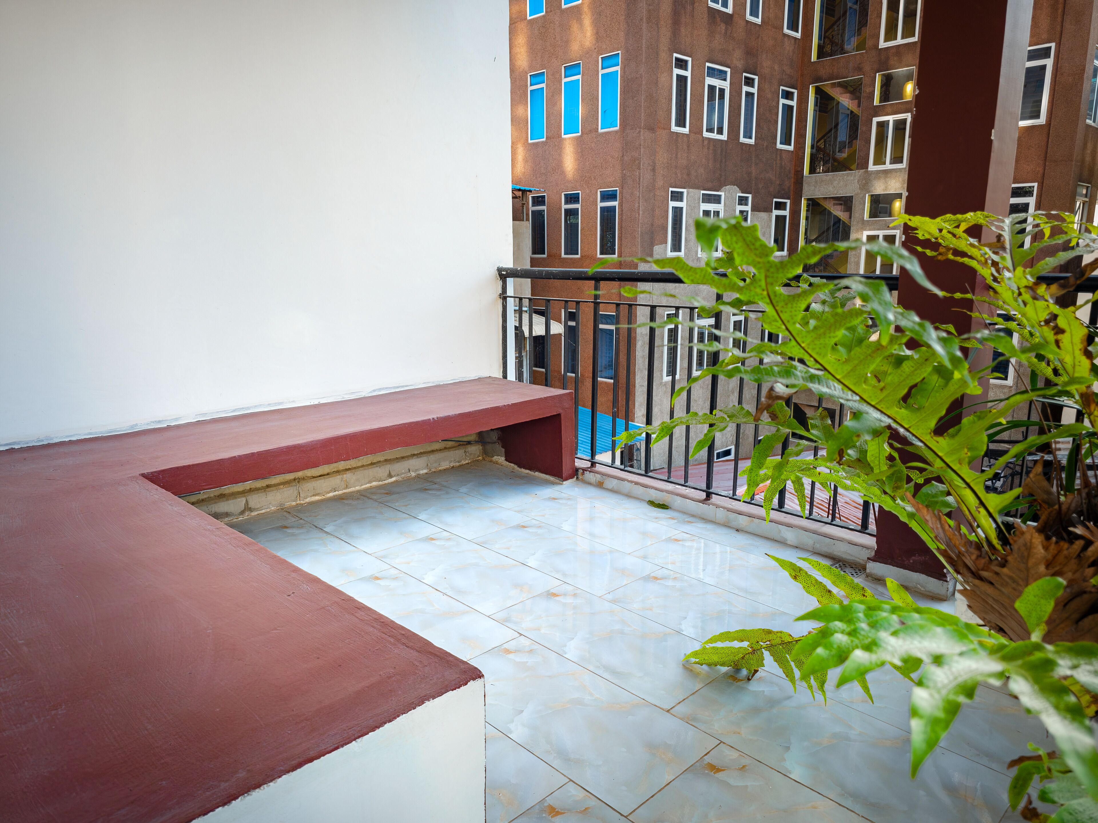 A balcony with a red bench, tiled floor, and a view of a multi-story building with blue windows.