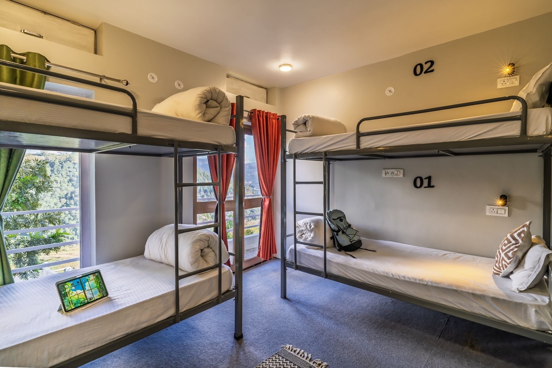 Une chambre de dortoir avec des lits superposés, numérotés 01 et 02, une tablette posée sur le lit du bas, et une vue sur la verdure à l’extérieur.