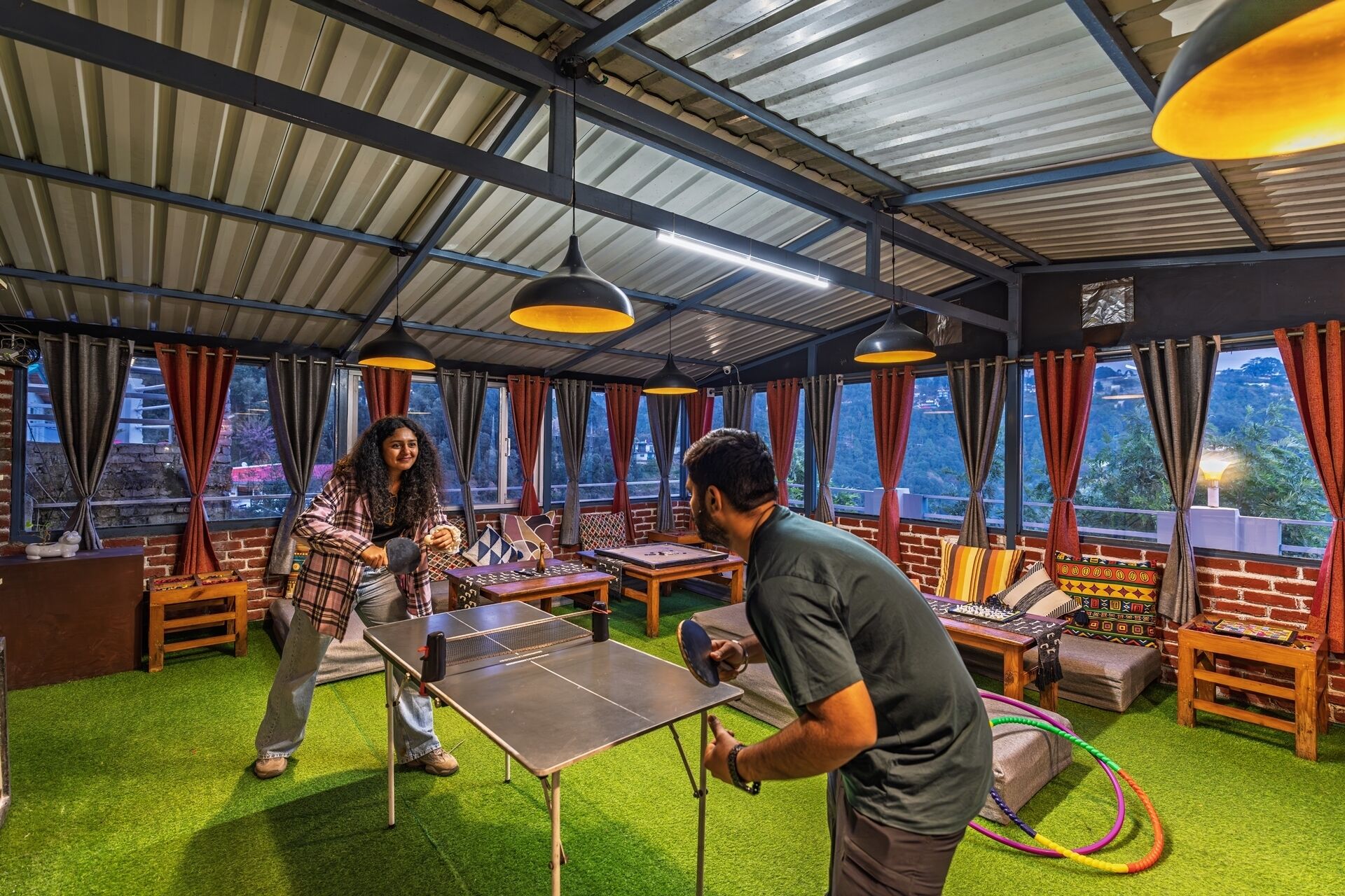 A room with a ping pong table, a brick wall, and a view of a mountainous landscape through large windows.