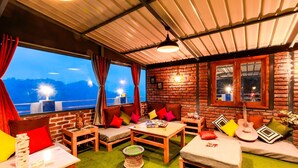 A cozy room with a brick wall, a wooden sofa, a small table, and a view of the mountains. - PACK Mukteshwar (Kosiyakutoli)