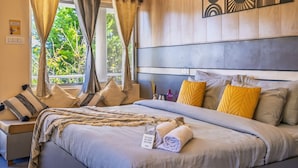 A well-lit bedroom with a large bed, multiple pillows, a bedside table with a lamp, and a view of greenery outside. - PACK Mukteshwar (Kosiyakutoli)