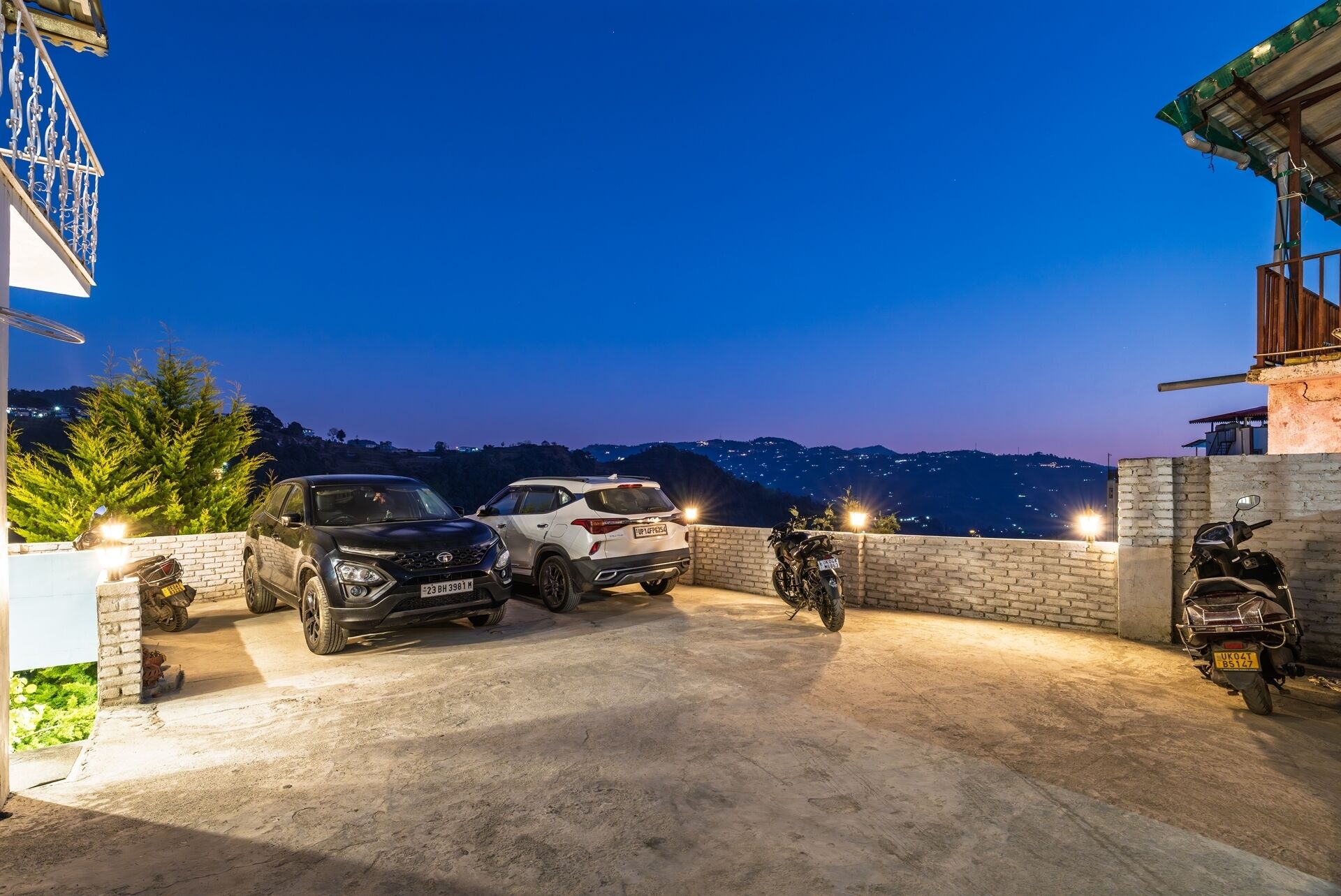 A parking area with two cars and a motorcycle, a brick wall, and a view of a hill with lights in the distance.