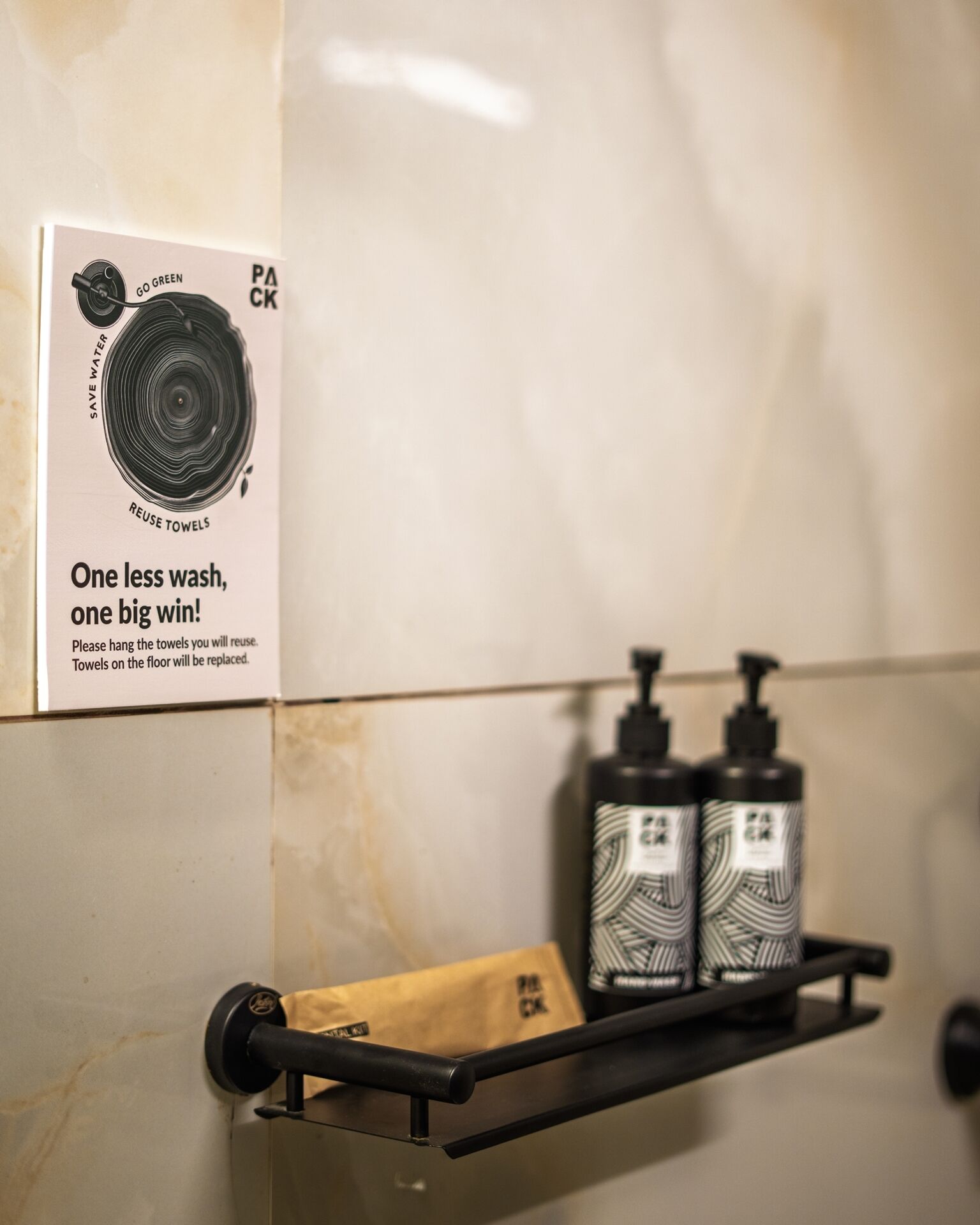 Une salle de bain avec un porte-serviettes sur lequel est posée une serviette marron, deux flacons de produits de toilette et un panneau encourageant la réutilisation des serviettes.