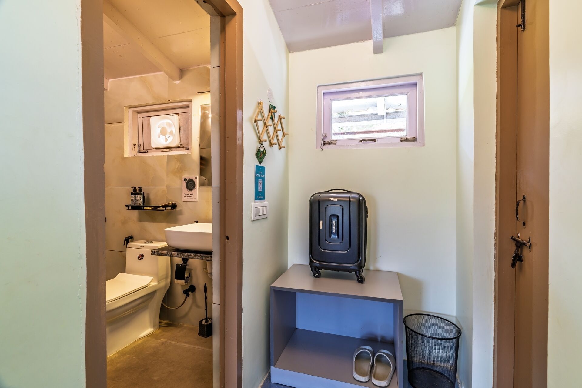 Ein kleines Badezimmer mit Toilette, Waschbecken und einem kleinen Fenster. Auf einem Regal steht ein Koffer und am Boden ein Paar Schuhe.