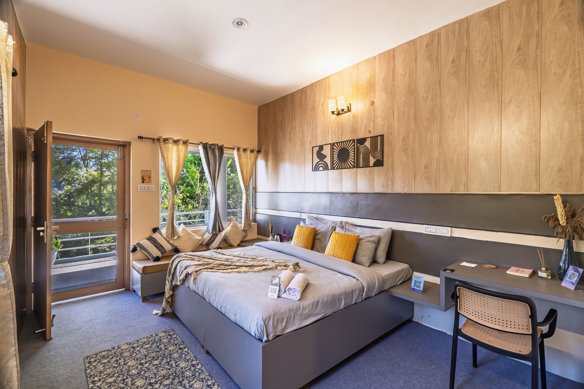 A modern bedroom with a large bed, a desk, and a chair. There is a window with curtains and a view of greenery outside.