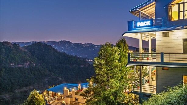 A hotel with a 'PACK' sign, a balcony with outdoor seating, and a view of a mountainous landscape. - PACK Mukteshwar (Kosiyakutoli)