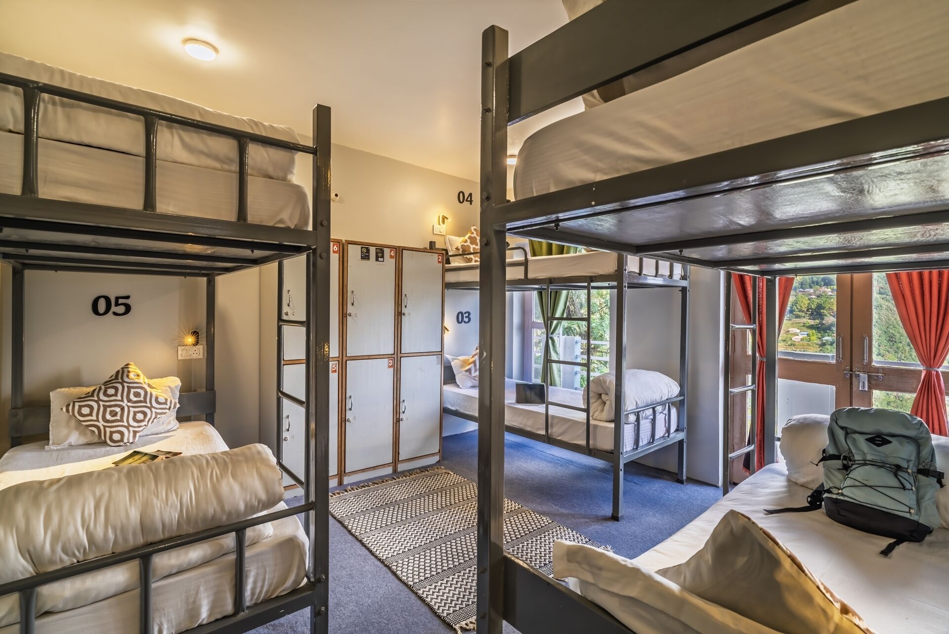 A dormitory-style room with bunk beds, numbered lockers, and a view of greenery outside.