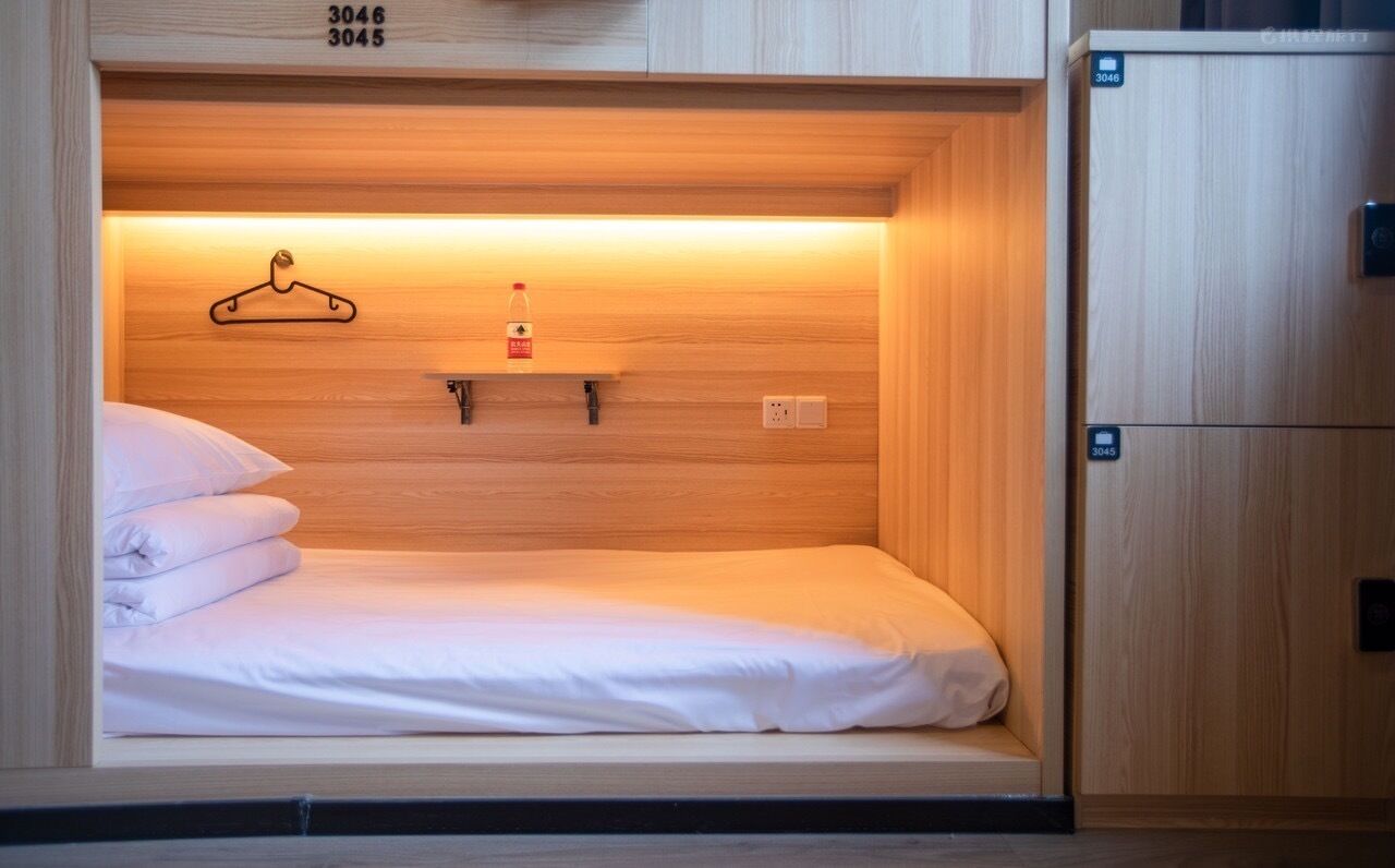 A compact hotel room with a single bed, a wooden headboard, a small shelf with a hanger and a bottle, and a wooden wardrobe with a lock.