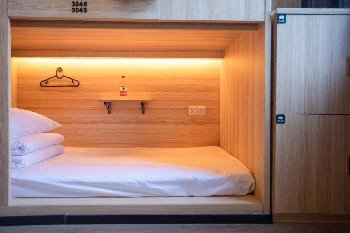Ein kleines Hotelzimmer mit einem Einzelbett, einem Holz-Kopfteil, einer kleinen Ablage mit Kleiderbügel und Flasche sowie einem abschließbaren Holzschrank.