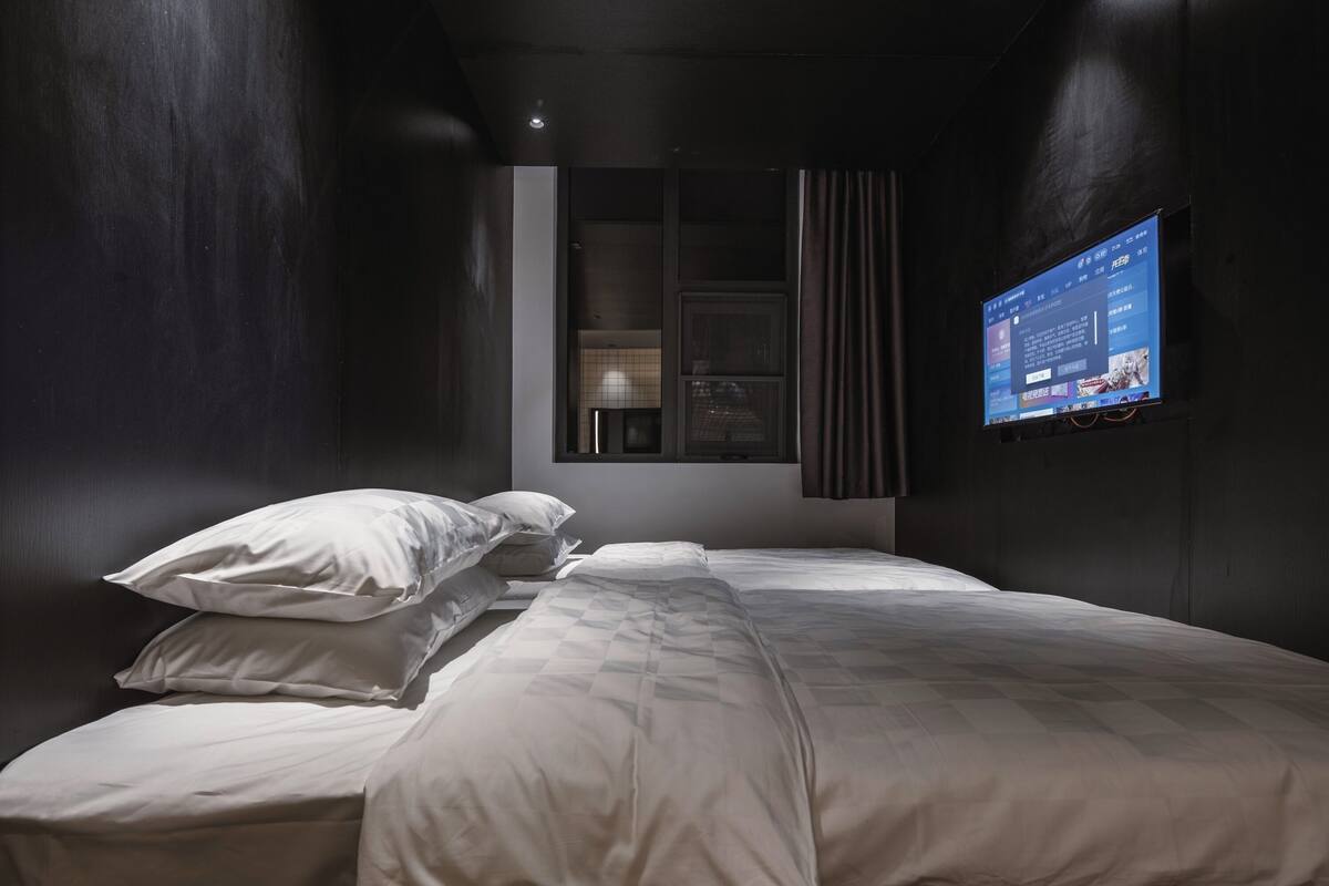 Ein modernes Schlafzimmer mit einem Bett, Kissen, einem an der Wand montierten Fernseher und einem Fenster mit Vorhängen.