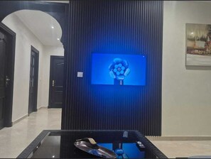 Un intérieur moderne avec un téléviseur à écran plat fixé sur un mur sombre, une table en verre avec une télécommande et une porte donnant sur une autre pièce.