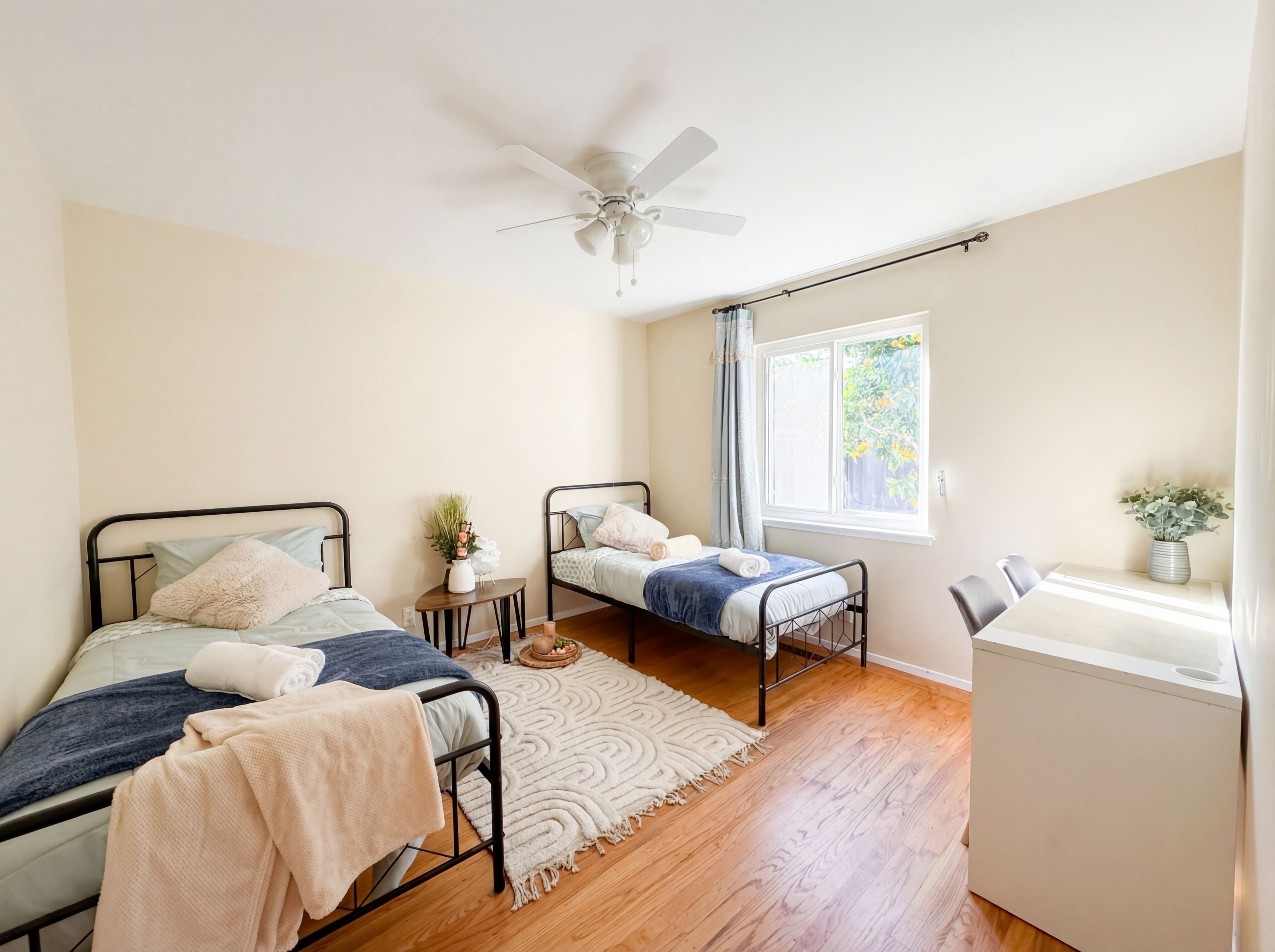 A bedroom with two beds, a ceiling fan, a window with a view of greenery, a small table with a plant, and a white radiator.