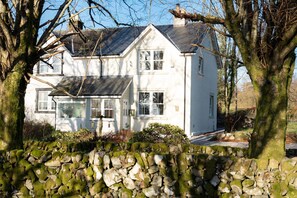 Une maison à deux étages, avec un mur de pierre à l’avant, entourée d’arbres.