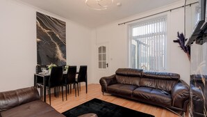 Una sala de estar moderna con un sofá de cuero marrón, un comedor con una mesa y sillas de vidrio, y un cuadro abstracto grande en la pared.