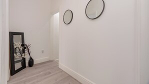 Un pasillo con paredes blancas y suelo de madera. Hay dos espejos redondos en la pared y un florero con flores en el suelo.