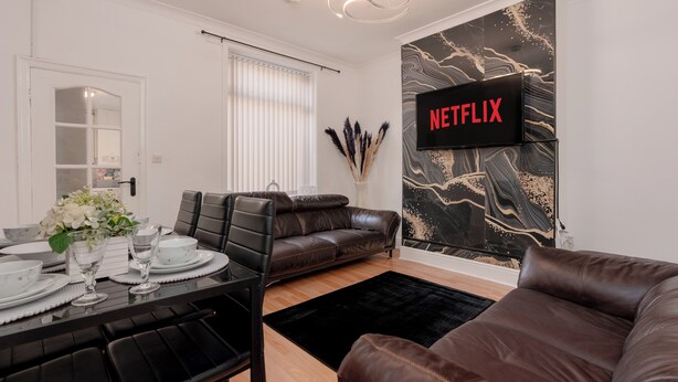 Una sala de estar moderna con un sofá de cuero marrón, una mesa de comedor con platos y vasos, un tapete negro y un televisor montado en la pared mostrando 