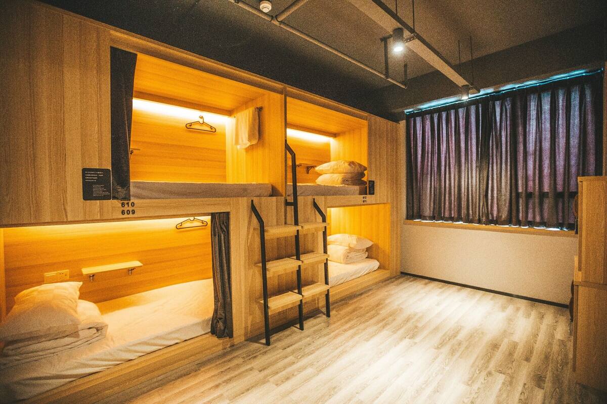Une chambre d’hôtel compacte avec des lits superposés, des murs en bois, une fenêtre avec des rideaux et un plancher en bois.