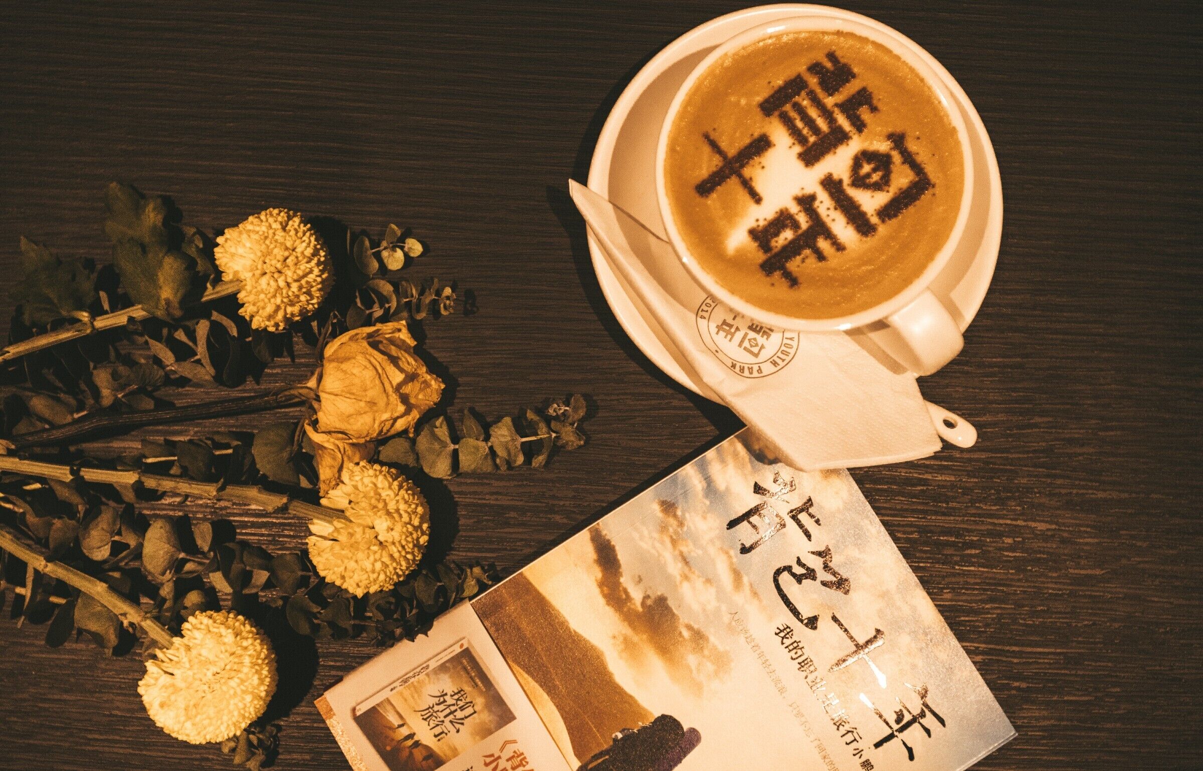 A cup of coffee with Chinese characters, a book, and dried flowers on a wooden surface.