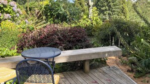 Una terraza de madera con una mesa y sillas de metal, rodeada de abundante vegetación y vistas a un lago.