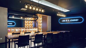 Un espacio moderno de bar con una larga mesa de madera, sillas negras y estanterías iluminadas que exhiben libros y revistas. Un letrero de neón en la pared dice "LIFE IS A JOURNEY".