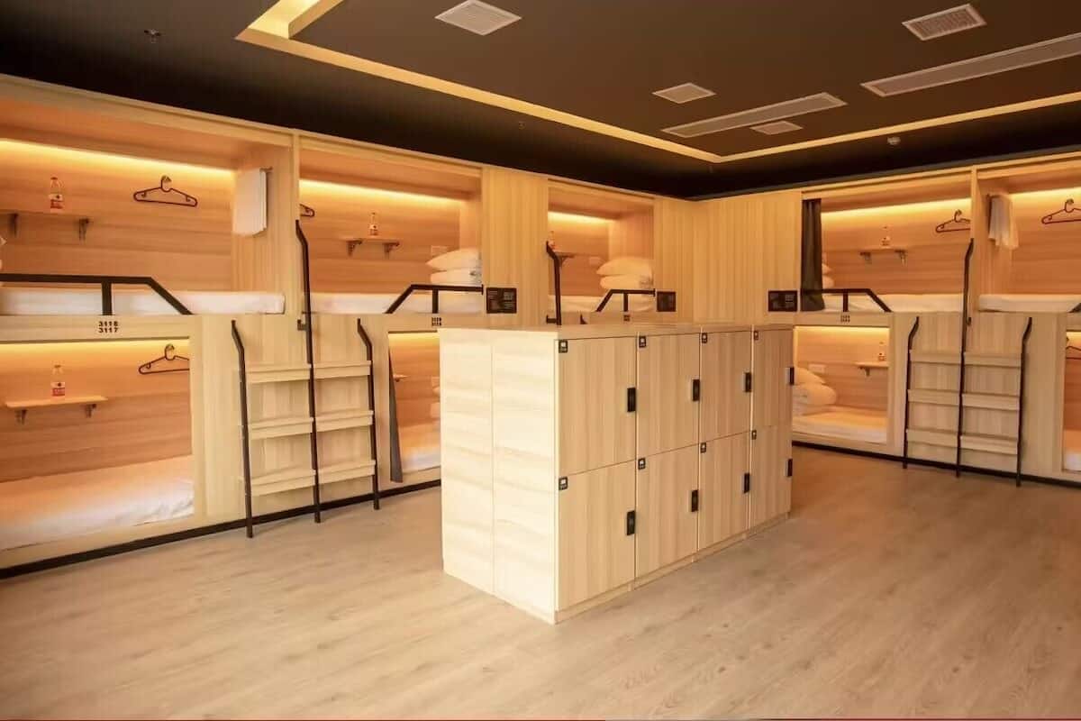 A modern hotel room with wooden paneling, a central wooden cabinet with multiple compartments, and a hanging rack with clothes.
