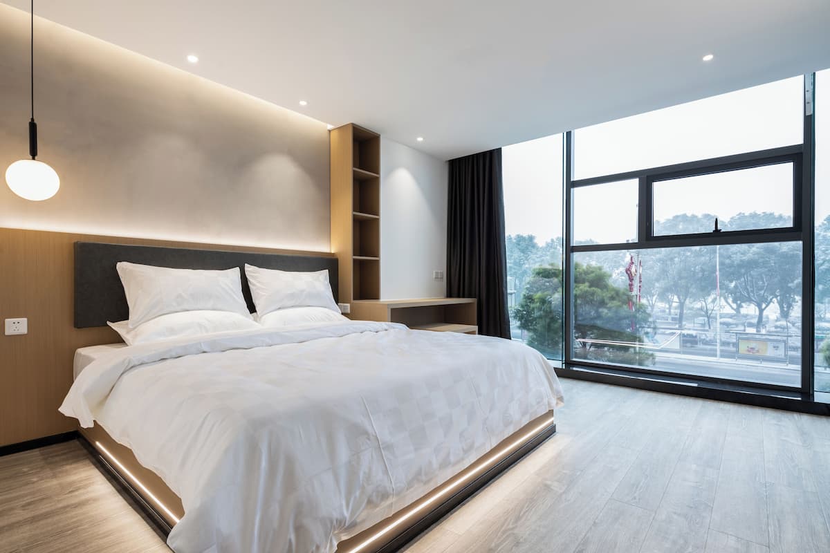 A modern bedroom with a large bed, built-in shelving, and a view of the outdoors through large windows.