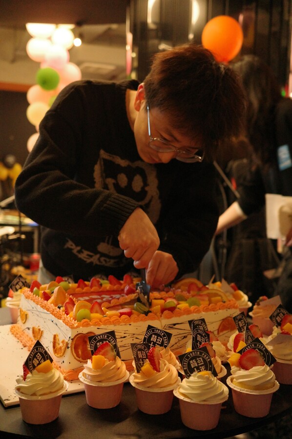 Une personne est en train de découper un gâteau à plusieurs couches, garni de fruits, et des cupcakes également décorés de fruits, le tout sur une table.