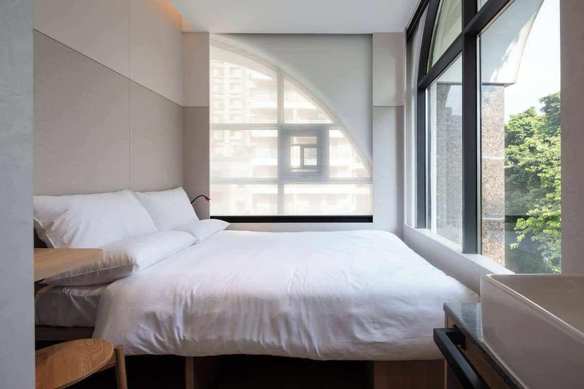 A modern bedroom with a large bed, a small desk, and a window with a view of buildings and trees.