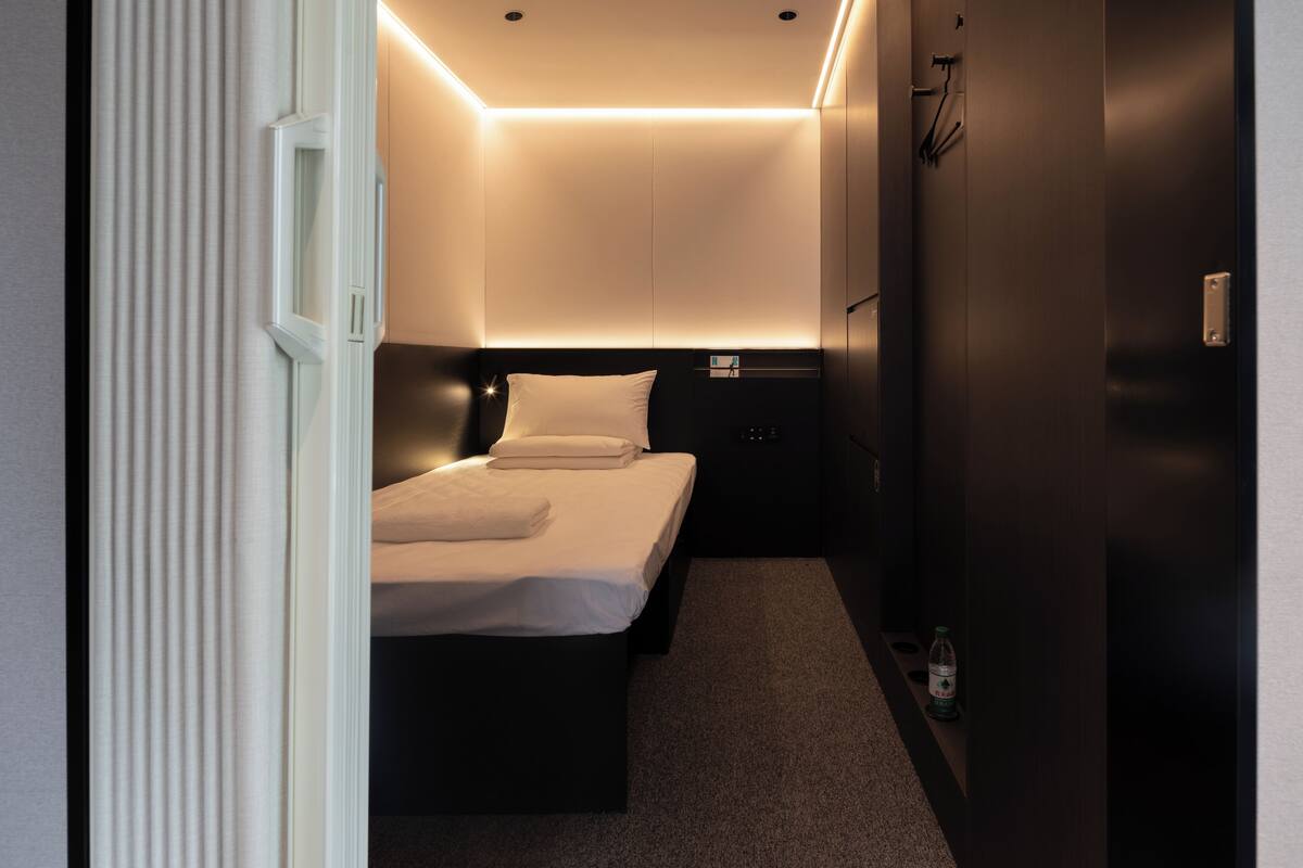 A compact hotel room with a single bed, a bedside table, a water bottle, and a wall-mounted TV.