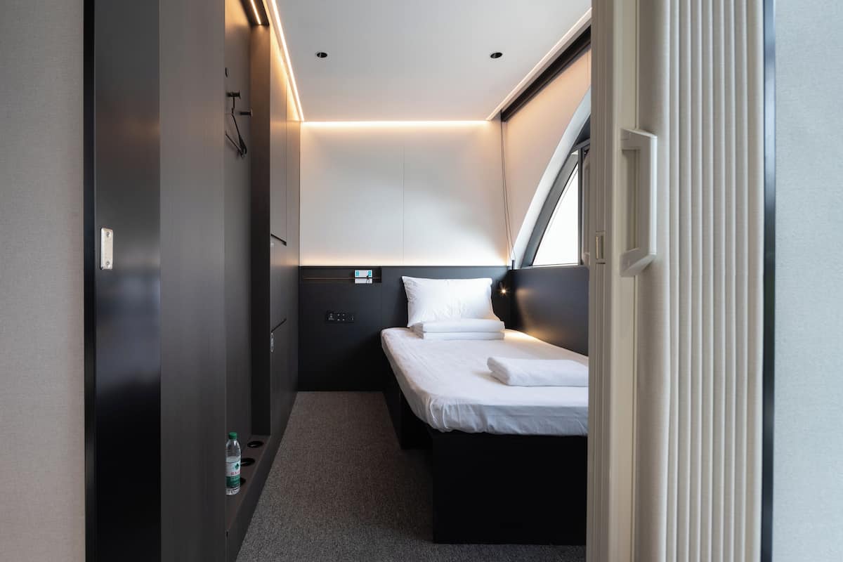 A compact, modern hotel room with a single bed, a bedside table, a small shelf, and a window with a view.