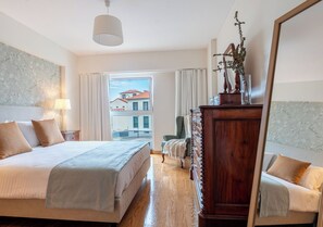 Ein Hotelzimmer mit einem großen Bett, einer Holzkommode, einem Spiegel und Blick auf Gebäude durch das Fenster.