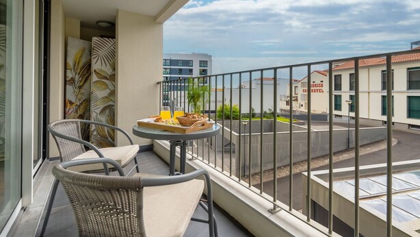 Ein Balkon mit Korbstühlen und einem Tisch, gedeckt mit Speisen, mit Blick auf eine Straße mit Gebäuden und blauem Himmel.
