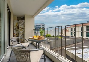 Ein Balkon mit Korbstühlen und einem Tisch, gedeckt mit Speisen, mit Blick auf eine Straße mit Gebäuden und blauem Himmel.