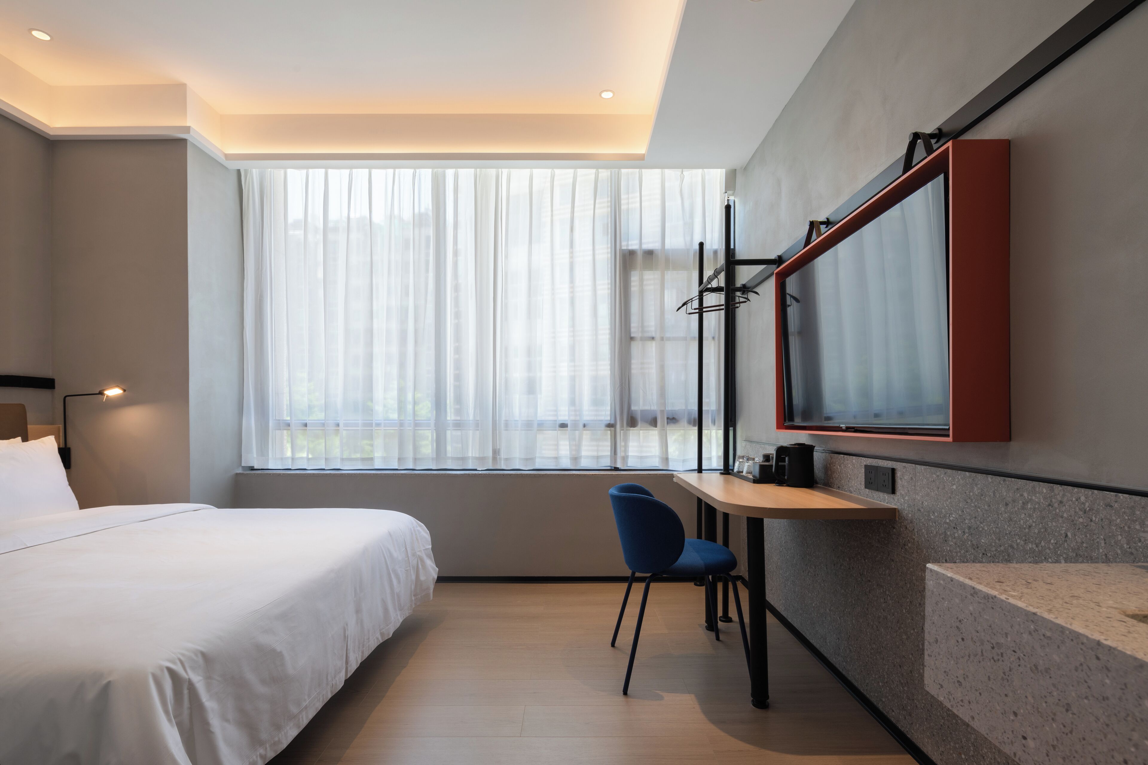 A modern hotel room with a bed, a desk, a chair, and a large window with sheer curtains.