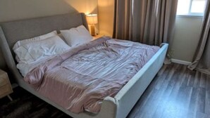 Een slaapkamer met een bed, een plafondventilator, een nachtkastje, een lamp, een raam met gordijnen en een houten vloer.