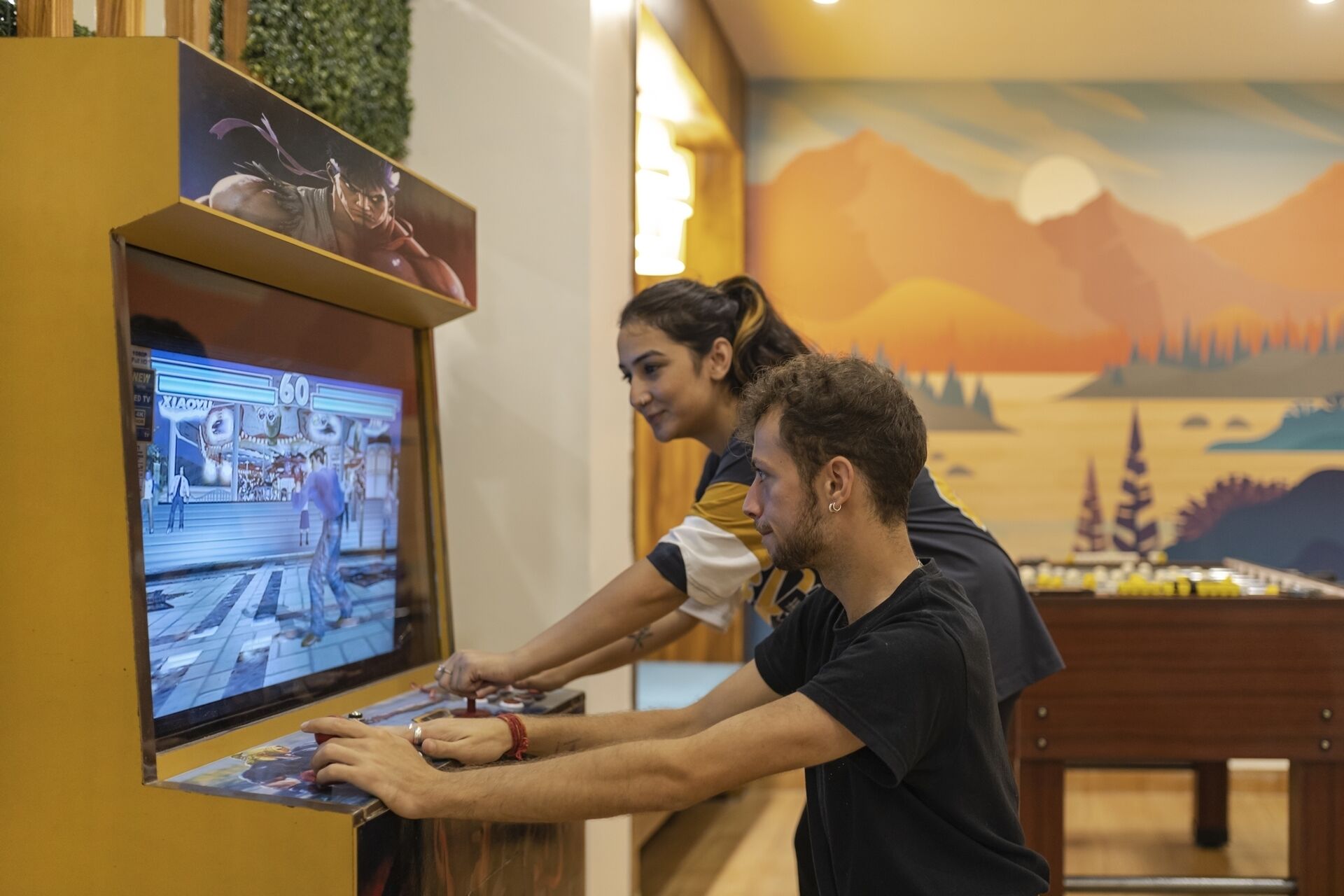 Dos personas jugando videojuegos en un quiosco, con un mural de fondo.