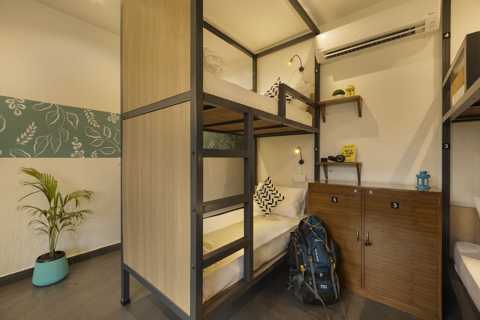 Habitación con literas, un armario de madera, un escritorio pequeño, una planta en maceta y un aire acondicionado instalado en la pared.