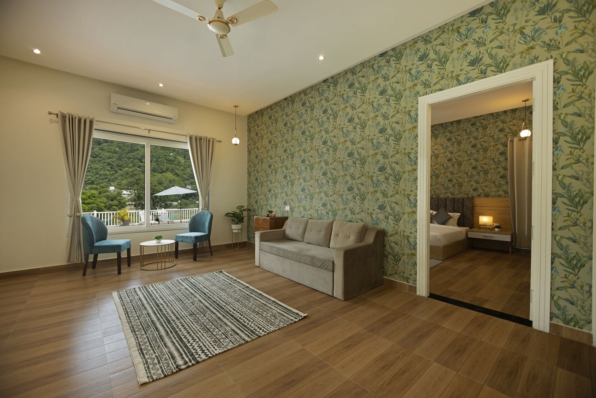 Amplia sala de estar con sofá, una mesita y una silla. Cuenta con ventana con cortinas, ventilador de techo y vistas al exterior con vegetación.