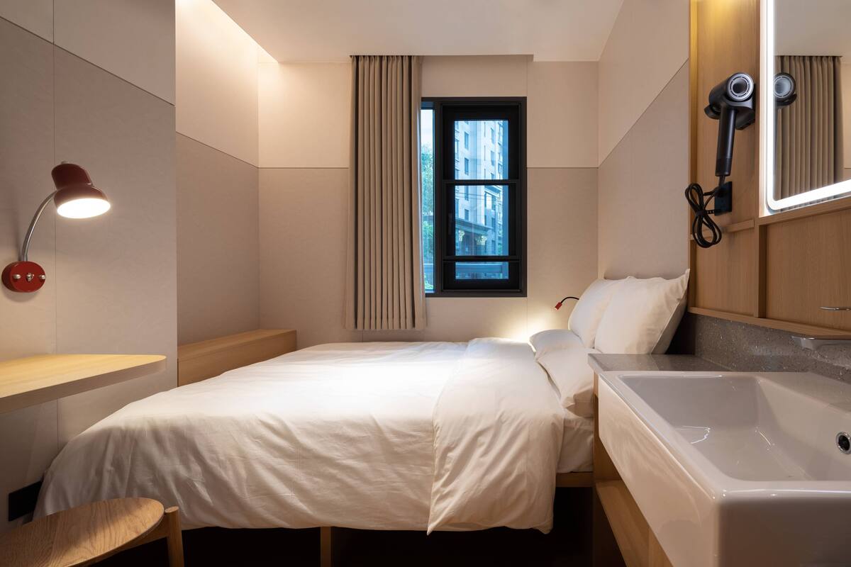 Een moderne hotelkamer met een bed, nachtkastje, bureau, stoel, raam met gordijnen, spiegel, haardroger en een bad.
