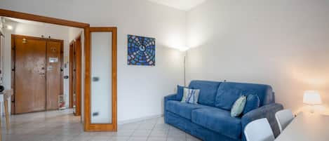Una habitación con un sofá azul, una mesa blanca, una lámpara, una obra de arte en la pared y una puerta que da a otra habitación.