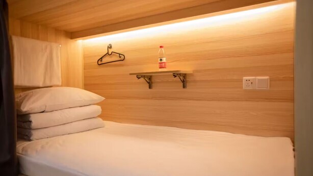 A single bed with a white mattress and pillows, a wooden wall, a shelf with a bottle, and a hanger. - Hangzhou Desti Youth Park Hostel West Lake (Hangzhou)