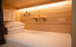Un lit simple avec un matelas et des oreillers blancs, un mur en bois, une étagère avec une bouteille et un cintre.
