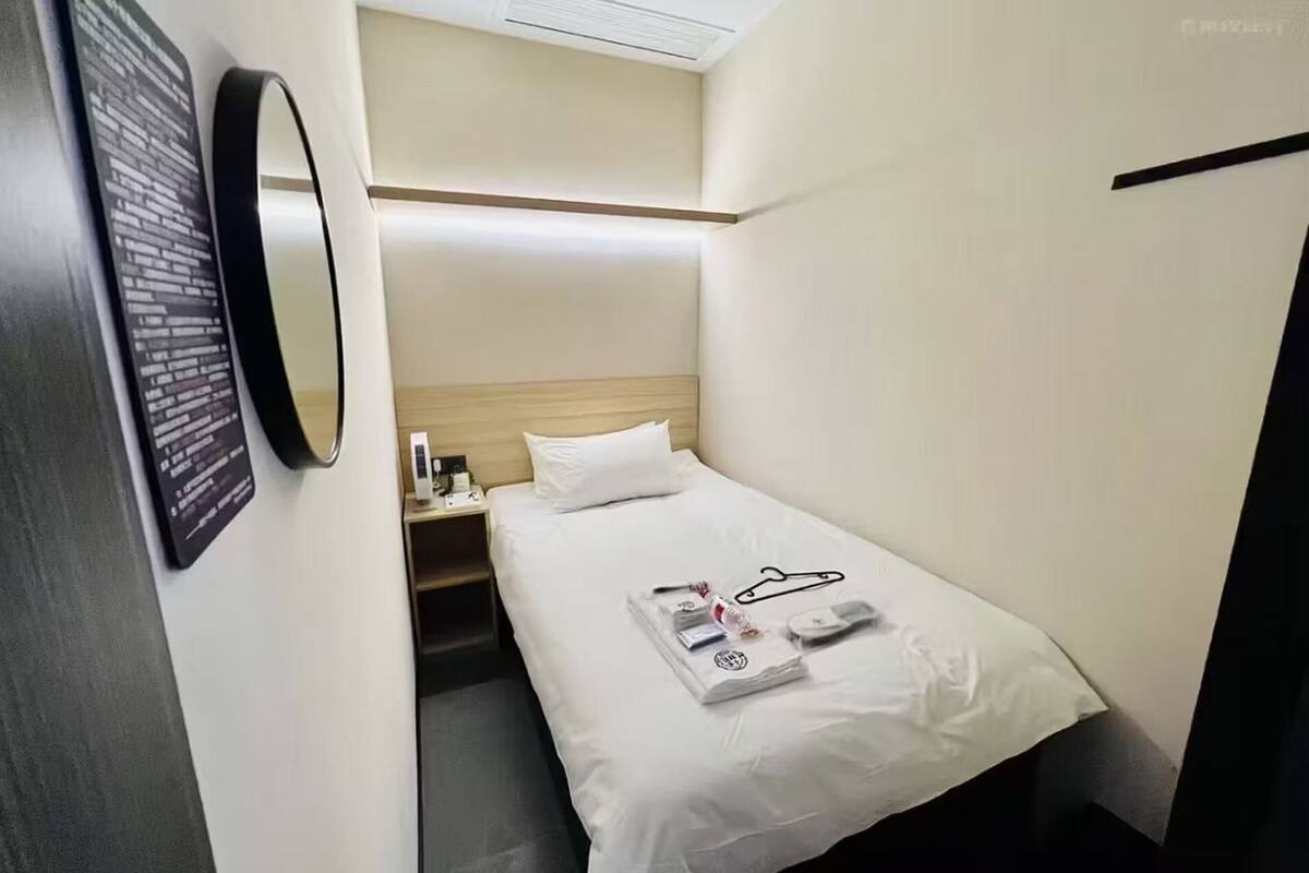 A hotel room with a single bed, a bedside table with a lamp, a mirror, and a wall-mounted information board.