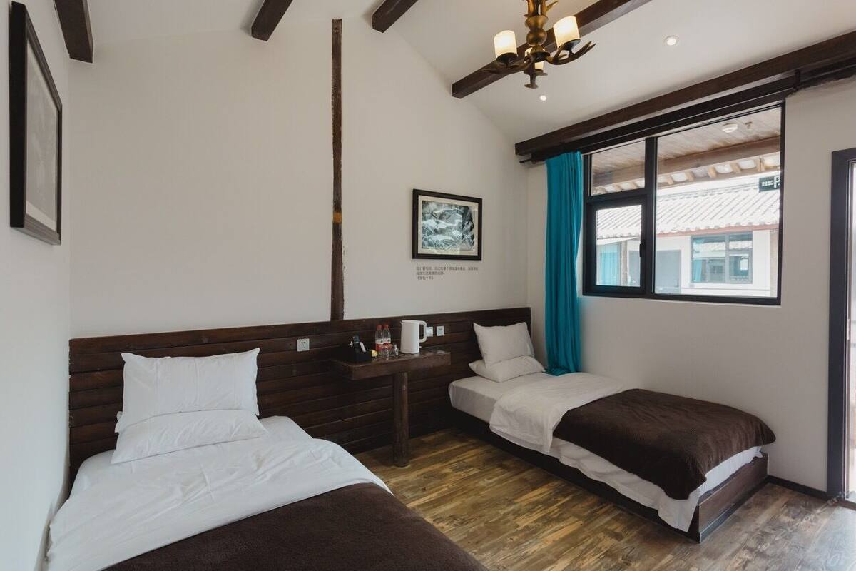 Ein Hotelzimmer mit zwei Betten, Holzbalken, einem Deckenventilator, einem Fenster mit Vorhängen und einem gerahmten Bild an der Wand.