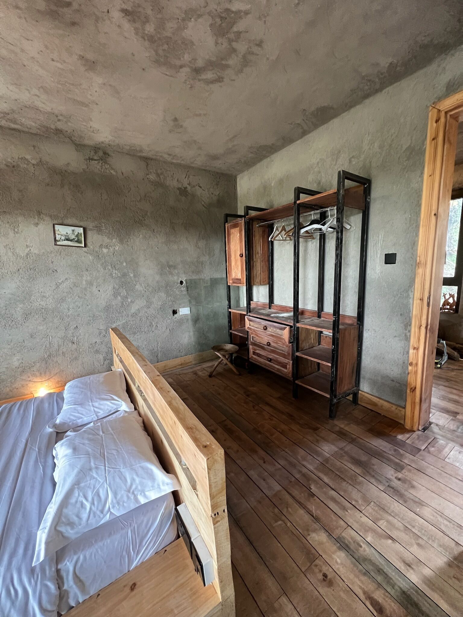 A room with a wooden bed, a wooden wardrobe with hanging space, and a concrete wall.