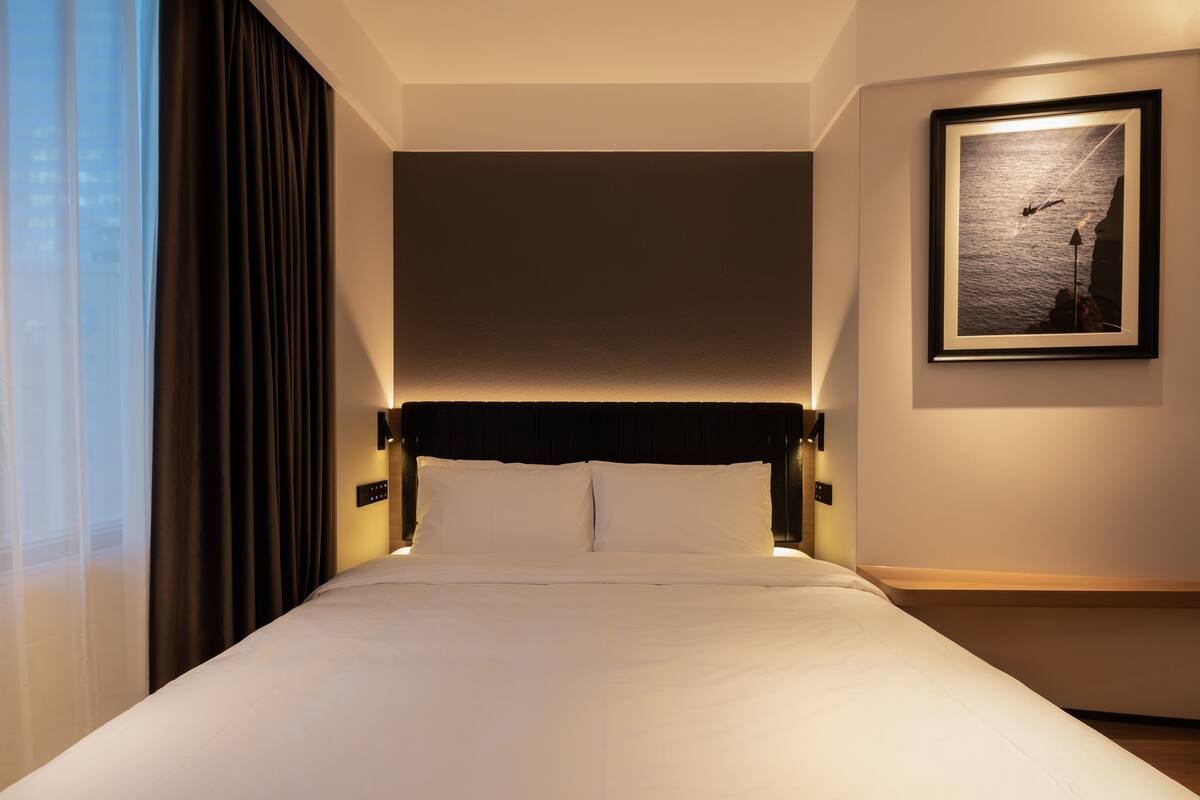 Quarto de hotel com cama bem arrumada, uma janela grande com cortinas, um quadro na parede e uma mesa de cabeceira com abajur.