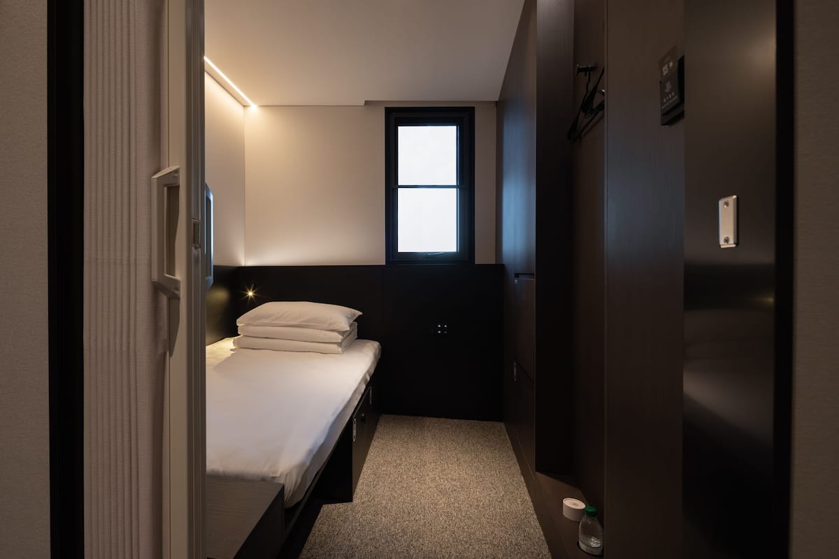 Quarto pequeno e com pouca luz, com uma cama, uma janela e uma porta.